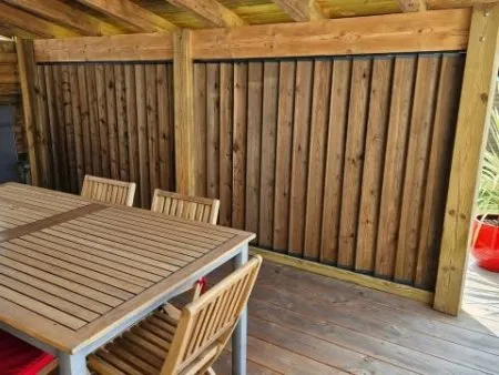 Terrasse en bois avec table et chaises, mur en lattes verticales, atmosphère chaleureuse et accueillante.