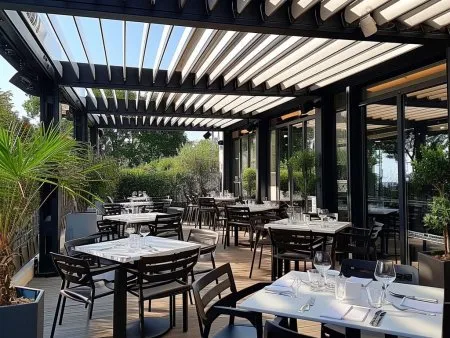Terrasse de restaurant moderne avec tables élégantes, pergola, verdure et vue sur l'extérieur, idéale pour un repas en plein air.