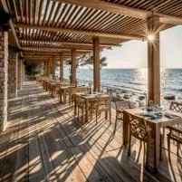 Terrasse d’un restaurant en bord de mer, tables en bois, lumière du soleil, ambiance calme et chaleureuse au coucher du soleil.
