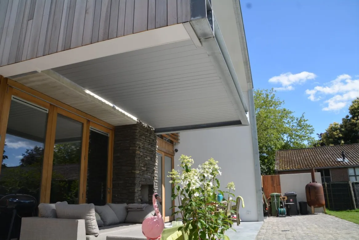 Terrasse moderne avec fauteuils confortables, fleurs, et façade en bois, sous un ciel bleu et ensoleillé.