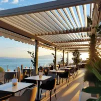 Terrasse moderne avec des tables blanches, chaises noires, et vue sur la mer au coucher du soleil, entourée de verdure.