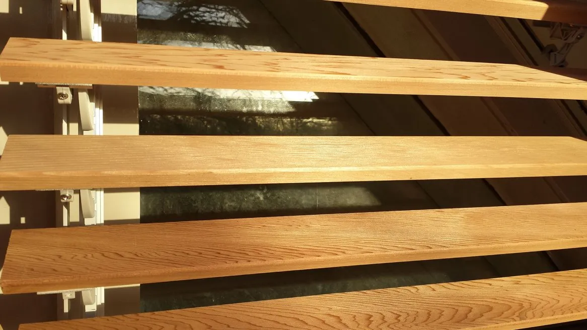 L’image montre des lattes en bois clair disposées horizontalement, laissant entrevoir un reflet derrière elles.