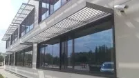 Immeuble moderne avec grandes fenêtres et stores. Vue sur un environnement verdoyant et un véhicule à l’arrière-plan.